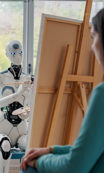 Robot humanoidalny malujący portret kobiety siedzącej naprzeciwko niego w jasnej, nowoczesnej pracowni artystycznej. Robot trzyma pędzel i paletę malarską, koncentrując się na płótnie umieszczonym na sztaludze. W tle widoczne są okna z widokiem na zieleń oraz akcesoria malarskie, takie jak ramy, pędzle i owoce ustawione na stole.