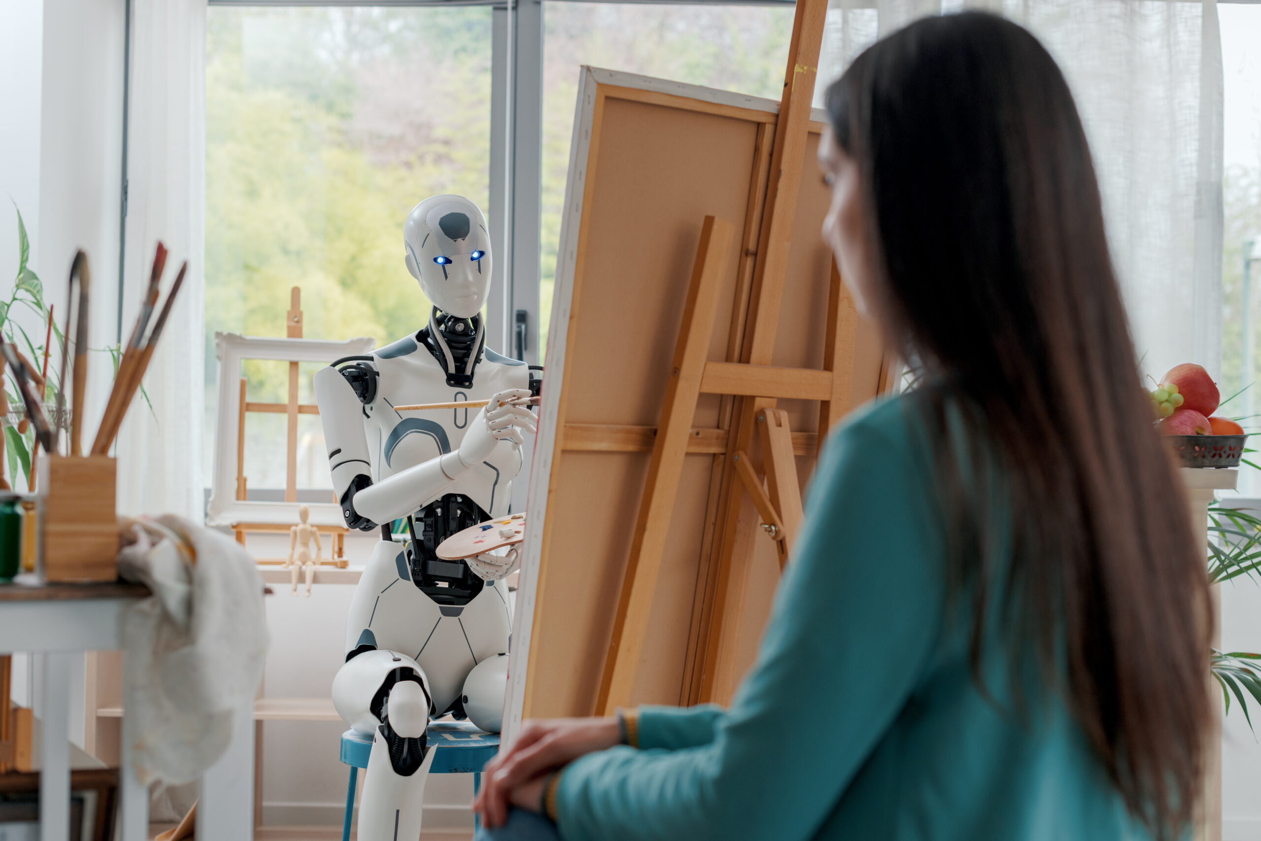 Robot humanoidalny malujący portret kobiety siedzącej naprzeciwko niego w jasnej, nowoczesnej pracowni artystycznej. Robot trzyma pędzel i paletę malarską, koncentrując się na płótnie umieszczonym na sztaludze. W tle widoczne są okna z widokiem na zieleń oraz akcesoria malarskie, takie jak ramy, pędzle i owoce ustawione na stole.